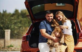 porta-malas está aberto. lindo casal tem um passeio junto com cachorro ao ar livre perto do carro foto