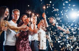 se divertindo com estrelinhas. confete está no ar. grupo de amigos alegres comemorando o ano novo dentro de casa com bebidas nas mãos foto