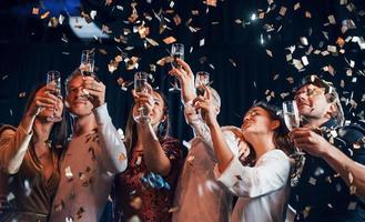 confete está no ar. grupo de amigos alegres comemorando o ano novo dentro de casa com bebidas nas mãos