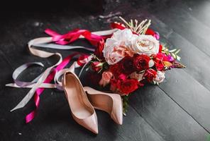 sapatos bege e buquê de noiva de rosas vermelhas em um piso de madeira preto. composição do casamento. foto