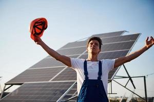 mãos levantadas. trabalhador masculino em uniforme azul ao ar livre com baterias solares em dia ensolarado foto