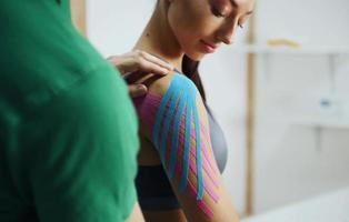 médico ajuda mulher por tratamento de ombro com fita kinesio foto