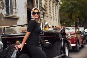 mulher loira de óculos escuros e vestido preto, apoiando-se no velho carro clássico vintage foto