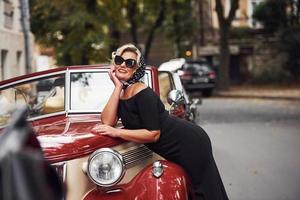 mulher loira de óculos escuros e vestido preto, apoiando-se no velho carro clássico vintage