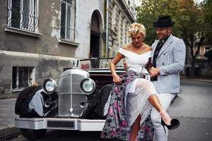 lindo casal com roupas antiquadas está na cidade com carro retrô foto