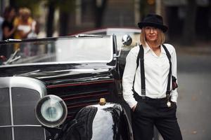 mulher loira de chapéu e em roupas retrô pretas perto de carro antigo clássico vintage foto