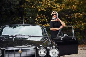 mulher loira de óculos escuros e vestido preto perto de carro antigo clássico vintage