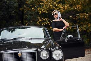mulher loira de óculos escuros e vestido preto perto de carro antigo clássico vintage foto