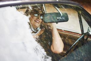 mulher loira de óculos escuros e vestido preto senta-se no velho carro clássico vintage foto