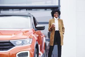 jovem afro-americana em copos e com copo de bebida fica ao ar livre perto de carro moderno foto