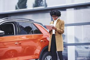 jovem afro-americana em copos e com copo de bebida fica ao ar livre perto de carro moderno foto