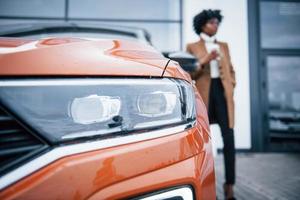 com copo de bebida. jovem afro-americana de óculos fica ao ar livre perto de carro moderno foto