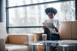 jovem afro-americana de óculos senta-se dentro de casa no escritório com o tablet nas mãos foto