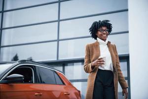 com copo de bebida. jovem afro-americana de óculos fica ao ar livre perto de carro moderno foto