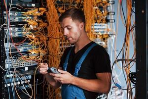 jovem de uniforme com dispositivo de medição trabalha com equipamentos de internet e fios na sala do servidor foto