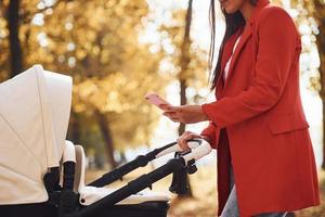 com telefone na mão. mãe de casaco vermelho dá um passeio com seu filho no carrinho no parque no outono foto
