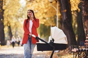 falando ao telefone. mãe de casaco vermelho dá um passeio com seu filho no carrinho no parque no outono foto