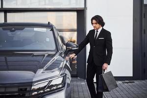 retrato do jovem empresário bonito de terno preto e gravata ao ar livre perto de carro moderno e com sacolas de compras foto