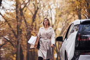 menina caminha perto do carro com sacolas de compras nas mãos. automóvel novo e moderno na floresta foto