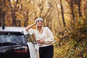 com copo de bebida nas mãos. menina tem viagem de outono de carro. automóvel novo e moderno na floresta foto