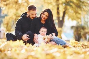 família alegre senta-se no chão e se diverte junto com seu filho no belo parque de outono