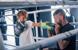 tendo sparring uns com os outros no ringue de boxe. jovem treinador de boxe tatuado ensina menina bonitinha no ginásio foto