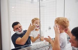 pai com seu filho está no banheiro se divertindo usando gel de barbear e olhando no espelho foto