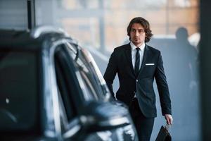 retrato do jovem empresário bonito de terno preto e gravata e com sacola de compras perto do carro moderno foto