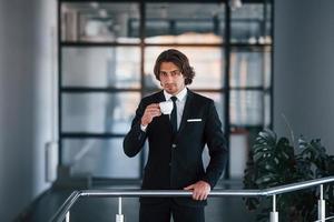 retrato de belo jovem empresário de terno preto e gravata fica dentro de casa com um copo de bebida