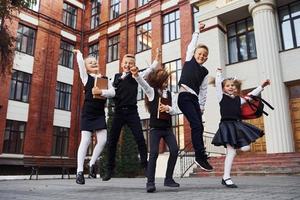 grupo de crianças em uniforme escolar pulando e se divertindo ao ar livre juntos perto do prédio da educação foto