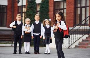 grupo de crianças em uniforme escolar posando para a câmera ao ar livre juntos perto do prédio da educação foto