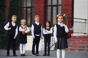 grupo de crianças em uniforme escolar posando para a câmera ao ar livre juntos perto do prédio da educação foto