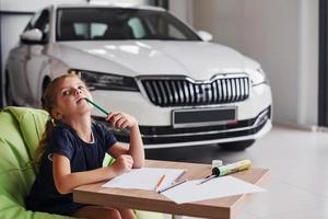 menina bonitinha senta-se na cadeira verde macia ao lado da mesa com lápis e folhas de papel. perto de automóvel moderno foto