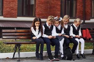 grupo de crianças em uniforme escolar senta-se no banco ao ar livre juntos perto do prédio da educação foto