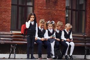 grupo de crianças em uniforme escolar senta-se no banco ao ar livre juntos perto do prédio da educação foto