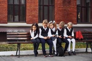 grupo de crianças em uniforme escolar senta-se no banco ao ar livre juntos perto do prédio da educação foto