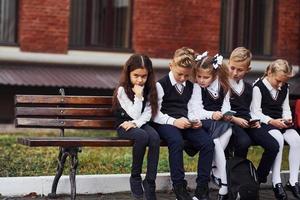 grupo de crianças em uniforme escolar senta-se no banco ao ar livre juntos perto do prédio da educação foto