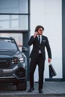 retrato de um jovem empresário bonito de terno preto e gravata ao ar livre perto de carro moderno foto
