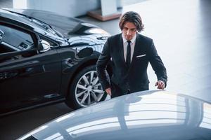 retrato de um jovem empresário bonito de terno preto e gravata dentro de casa perto de carro moderno foto