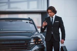 retrato do jovem empresário bonito de terno preto e gravata ao ar livre perto de carro moderno e com sacolas de compras foto