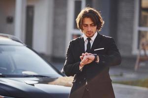 retrato do jovem empresário bonito de terno preto e gravata ao ar livre perto de carro moderno na cidade foto