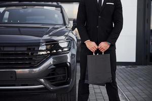 close-up vista do empresário de terno preto e gravata fica perto de automóvel moderno com sacolas de compras nas mãos foto
