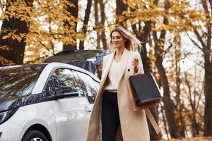 menina caminha perto do carro com sacolas de compras nas mãos. automóvel novo e moderno na floresta foto