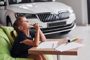 menina bonitinha senta-se na cadeira verde macia ao lado da mesa com lápis e folhas de papel. perto de automóvel moderno foto