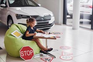 menina bonitinha senta-se na cadeira verde macia ao lado da mesa com lápis e folhas de papel. perto de automóveis modernos e sinais de trânsito no chão foto