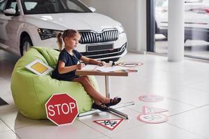 menina bonitinha senta-se na cadeira verde macia ao lado da mesa com lápis e folhas de papel. perto de automóveis modernos e sinais de trânsito no chão foto