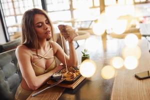mulher loira sexy com roupa quente senta-se à mesa e come fast-food dentro de casa no café foto