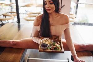 jovem morena em roupas sexy se estica sentando na mesa e segura o prato com comida no restaurante foto