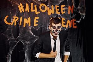 retrato do homem que está na festa temática de halloween em maquiagem de esqueleto assustador e fantasia contra a parede da cena do crime foto