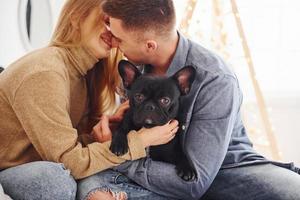 feliz jovem casal moderno sentado no sofá em casa com a árvore de natal com seu cachorro fofo foto